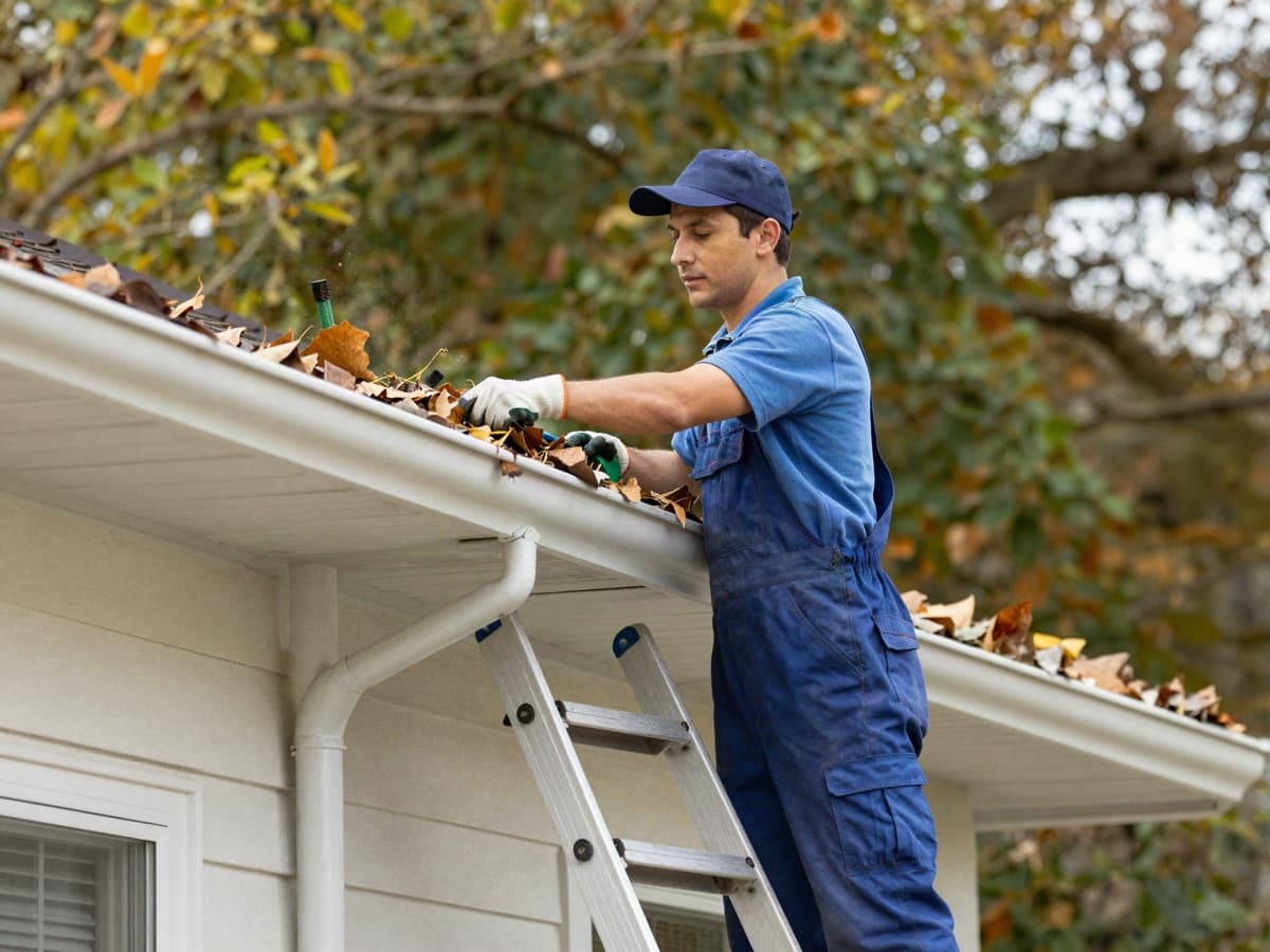 A worker on a ladder clearing fallen leaves from a house gutter along the roof eave