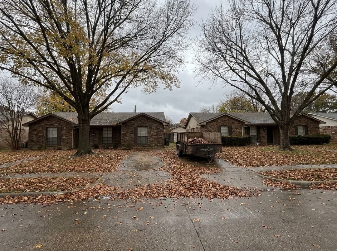 Two neighboring brick homes on a North Texas street in November, lawns buried in pecan and oak leaves, with one dump trailer parked between the driveways.