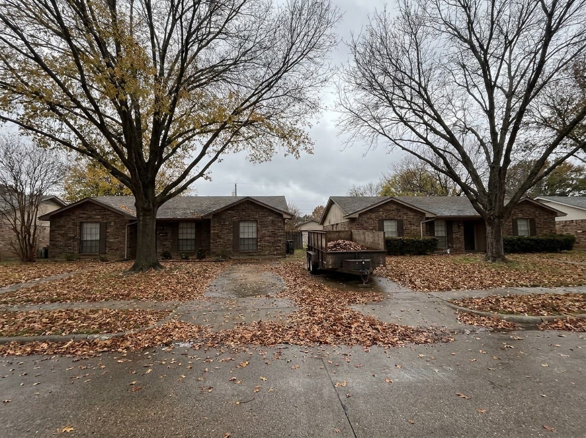 Two neighboring brick homes on a North Texas street in November, lawns buried in pecan and oak leaves, with one dump trailer parked between the driveways.